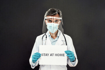 Mature male doctor in medical uniform, protective mask and face shield holding paper with text STAY AT HOME while standing against dark background. Selective focus on a man