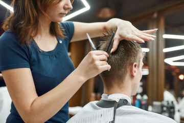 Professional barber girl holding scissors and comb in hands and making haircut for her client. Young guy visiting barbershop