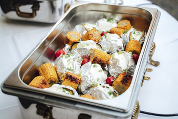 Grilled corn and baked potatoes stuffed with cheese in hotel pan on food warmer. Self-service buffet table. Celebration, party, birthday or wedding