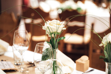 Closeup of festive table floral decoration. White roses on table with white table cloth. Table setting with cutlery and wineglasses. Celebration