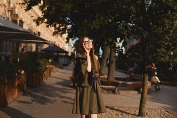 Fototapeta premium Portrait of a cheerful young woman in dress touching her sunglasses on urban background. Fashionable girl with beautiful brown hair smiling to camera.