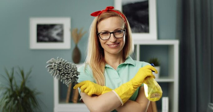 Portrait of beautiful mature housewife wearing eyeglasses and yellow rubber gloves, holding bottle with cleaning spray and rag, smiling and looking at camera.