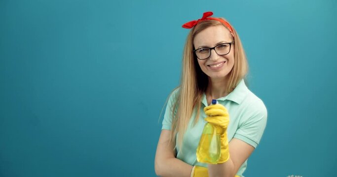 Charming woman in eyeglasses and rubber gloves holding in one hand cleansing spray and brush on another. Isolated over lue studio background. Concept of housekeeping.