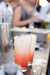 Two colorful summertime cocktails being prepared in tall glasses at the bar outside.