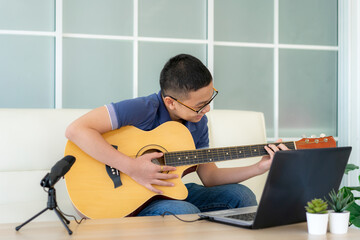 Asian boy playing acoustic guitar and watching online course on laptop while practicing at home. Asian boy virtual happy hour meeting for play music online together with friend via videocall.