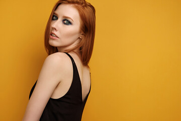 Red-haired girl in a black top and with blue eye make-up stands on a yellow background, looking into the frame over her shoulder