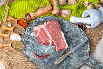 Raw t bone steak is on a wooden Board on black background, next to salt, pepper and herbs.