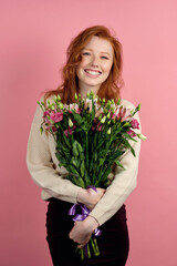Cute red-haired girl stands on a pink background, hugging a bouq