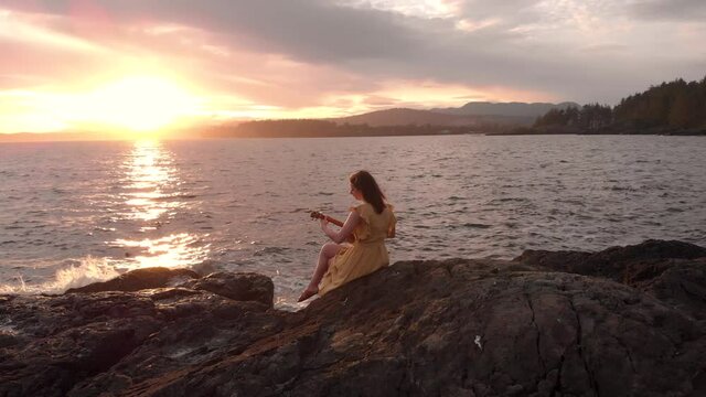 Young attractive brunette woman playing a ukulele by the ocean at sunset. 4k 24fps.