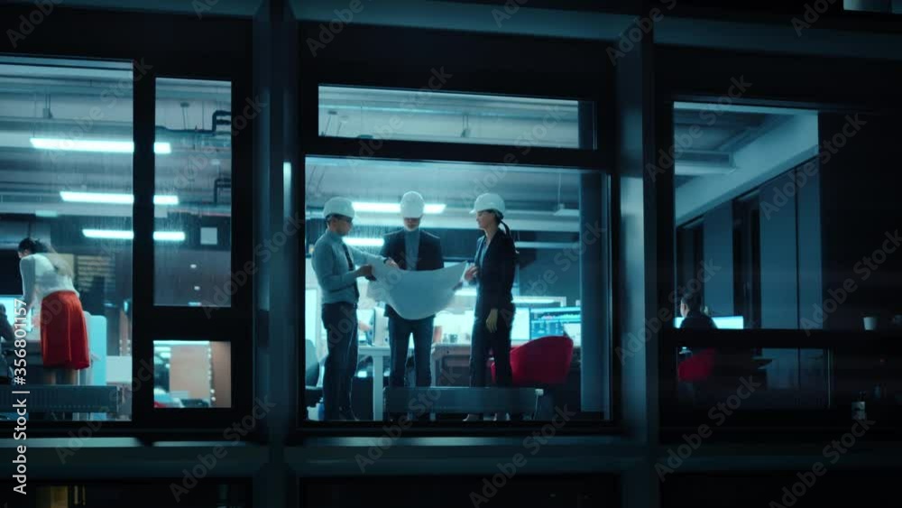 Poster Footage from Outside the Window: Architects and Engineers in Hard Hats Stand Next to a Window in Office Workplace and Look at Blueprints on Paper. Senior Specialists Explains the Job to His Apprentice