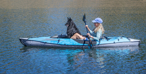 A blonde woman with a black Belgian Sheepdog dog paddling an inflatable kayak and looking forward on a blue lake © Janet
