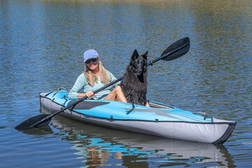 A smiling blonde woman in a kayak with a black Belgian Sheepdog dog paddling toward camera and looking in to camera on a blue lake © Janet