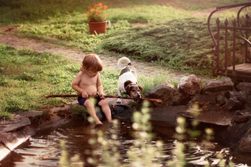 little boy 2 years old sits on shore of pond in village in his hand stick, next to friend is dog Jack Russell