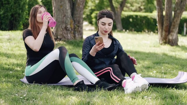Pretty sporty women are sitting at the park. The ladies are having a break from training.