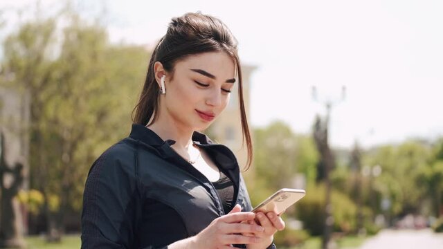 An athletic woman is standing outside setting her smartphone. She is using earpods and wearing sportswear.
