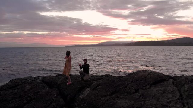 A attractive young man getting on his knee to propose to his girlfriend beside the ocean at sunset. 4k 24fps.