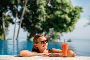 Happy smiling woman win sunglasses with orange juice in the swimming pool. Beautiful lady drinking a cocktail in a swimming pool and relaxing