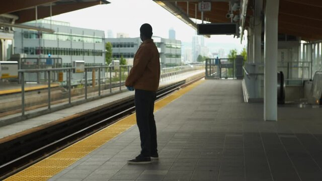 African American black man waiting for train on exterior platform wearing causal clothes and PPE mask and gloves. 4K.