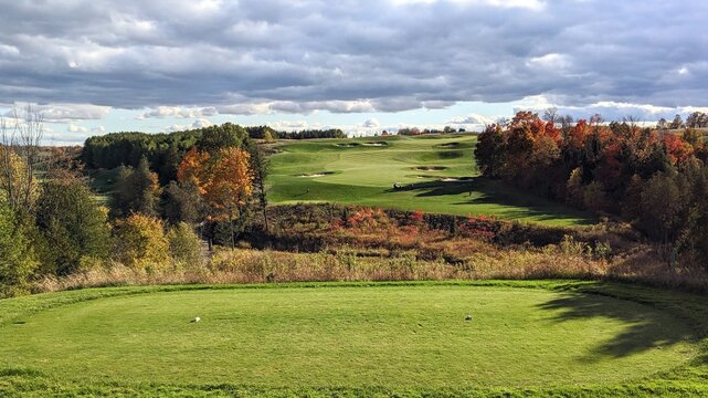 Views of a golf course hole from the tee box.  Straight ahead is a clean fairway with sand bunkers on both sides.  On the side of the rolling hills are trees changing color, under a cloudy backdrop.