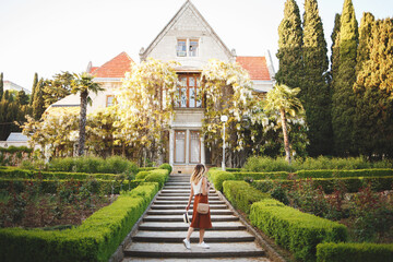 Young beautiful woman, wearing skirt, top and hat, walking in a blooming park among the greenery and the old palace with wisteria.