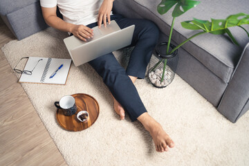 Close up of young Asian man sitting on floor working from home using laptop computer and notebook. Work from home concept