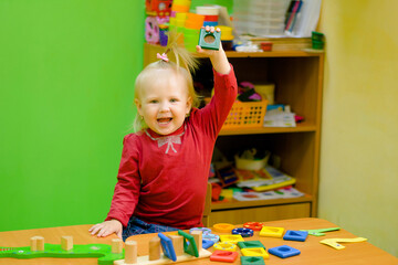 Little girl playing with toys - blocks. Kid girl with a raised hand at school for classes developing lessons for children. Colorful classroom.