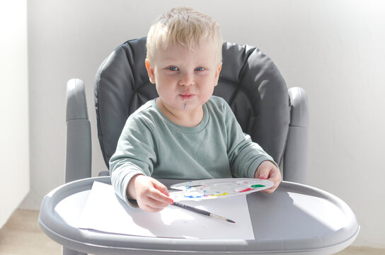 Blonde toddler boy painting with watercolor and brush on white paper in kids chair. Drawing Development In Children