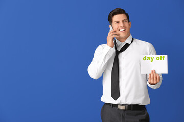 Young businessman holding paper with text DAY OFF while talking by phone against color background