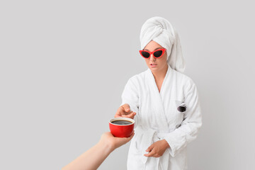 Hand offering cup of coffee to young woman in bathrobe on grey background
