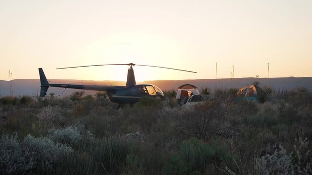 Campsite with tents and helicopter on top of desert mountain at sunset with sun flare as pilot walks towards cockpit slow motion