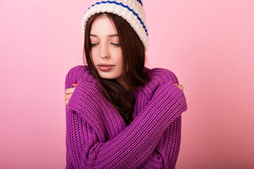 Cute young brunette girl in a knitted sweater and hat in winter headphones and mittens on a pink background.