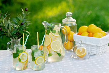 Lemonade refreshing drink in a jug and jars with lemons, fresh mint and ice with a basket with lemons and kumquat on a garden table. Summer outdoor picnic. Soft selective focus.