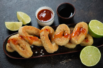 Fried breaded calamari rings with lime and dipping sauces, studio shot on a dark brown stone surface, close-up
