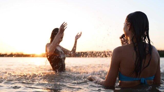 Happy wet girls in bikini run into sea and play splashing water to each other on sunset. Cheerful female friends have fun making splashes in pond. Young women go to swim, bathe in lake. Slow motion.