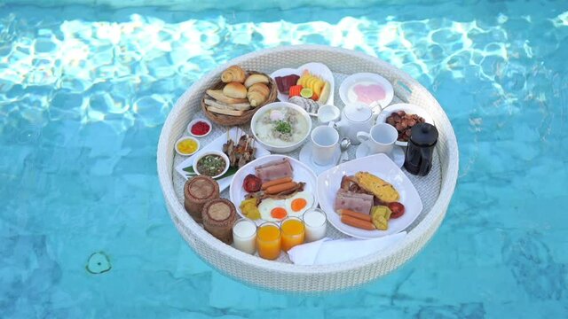 Top view on lunch food tray floating on the pool water. Coffee cups, glasses with juice, bread, fruits, eggs bacon of floating tray at hotel