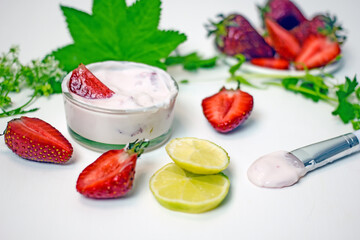 DIY homemade strawberry facial mask. A glass bowl with a pink mass of cream and strawberries next to a sprig of green plants, halves of fresh berries, lime, a brush for applying masks on a white backg