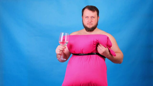 Funny bearded guy in a pink dress made of pillows drinks wine from a glass on a blue background. Crazy quarantine. Fashion 2020. Put on a pillow. Challenge 2020 due to house isolation