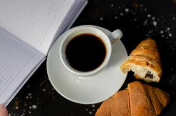 cup of hot coffee on black background with croissant and notebook