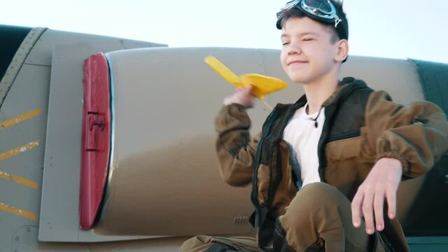 Happy dreamy smiling kid boy launches toy, thinking fantasy of flying sitting on wing of real airplane. Future little pilot plays costume adventure aviation game in profession. Joy, interest childhood