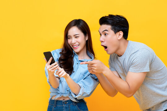 Amazed Attractive Young Asian Couple Looking At Smartphone Together Isolated On Yellow Studio Background