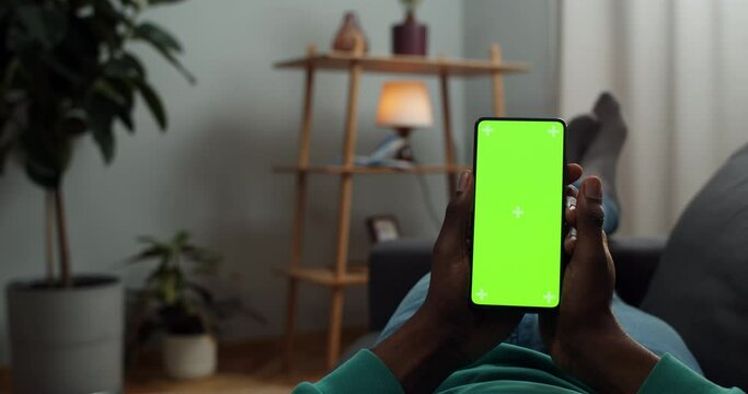 Young man lying on sofa with green screen and trecking markers smartphone in his hands. Guy reading news or e book while looking at mock up screen. Concept of chroma key. Cosy home background.