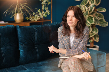 Beautiful happy woman brunette psychologist listens and speaks with a patient. Female psychotherapist consulting at office. Sitting on the sofa, makes notes.