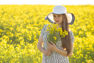 Girl in a rapeseed field, in a hat, with a bouquet of rapeseed in her hands.