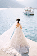 Fototapeta na wymiar The bride stands on the pier of the city of Perast in Montenegro, and admires the mountains and moored boats. The train of the dress is laid out on the floor and a long veil develops in the wind.
