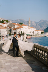 Fine-art wedding photo in Montenegro, Perast. A wedding couple on the hotel terrace with panoramic views of the old town of Perast, the groom circles the bride with a long dress on her hands.