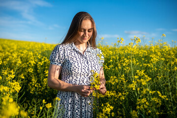 Naklejka na ściany i meble A girl in a rapeseed field, with her hair loose, collects a bouquet.