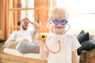 Portret of playful little daughter playing funny game as doctor having fun with dad sitting on couch, cute child girl pretending nurse holding stethoscope listening to father patient at home