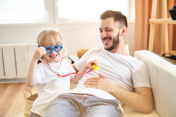 Playful little daughter playing funny game as doctor having fun with dad sitting on couch, cute child girl pretending nurse in medical uniform holding stethoscope listening to father patient at home