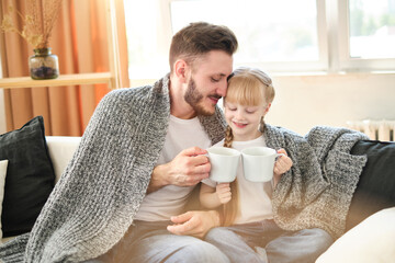 Father and daughter. Handsome young man and little cute girl sitting on the sofa at home, covered themselves with a warm blanket, holding white cups with hot tea on a sunny winter day. Father's day. - Powered by Adobe