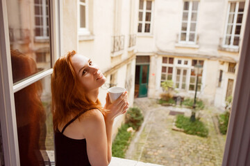 woman drinking coffee in morning by window overlooking inner courtyard. 